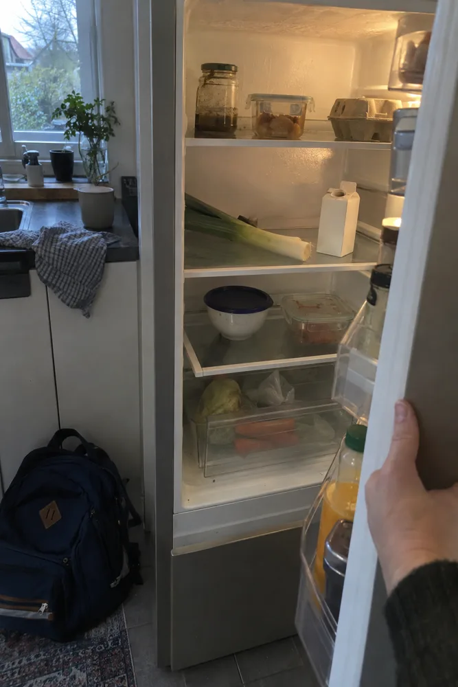 Een hand houdt om vijf uur de koelkastdeur open: een halve prei, een geopend pak room