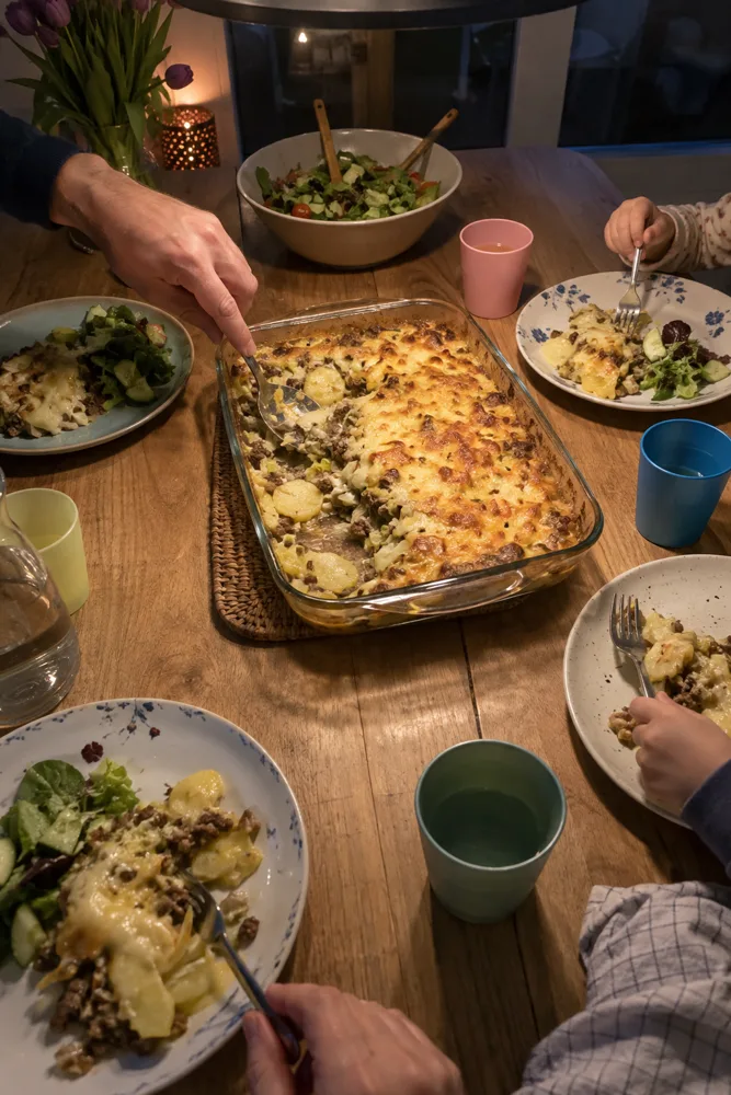 Om zes uur aan tafel: een ovenschotel met goudbruine kaas, vier borden en handen die opscheppen